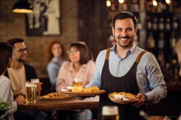 Garçom feliz segurando pratos com comida e olhando para a câmera enquanto serve convidados em um restaurante