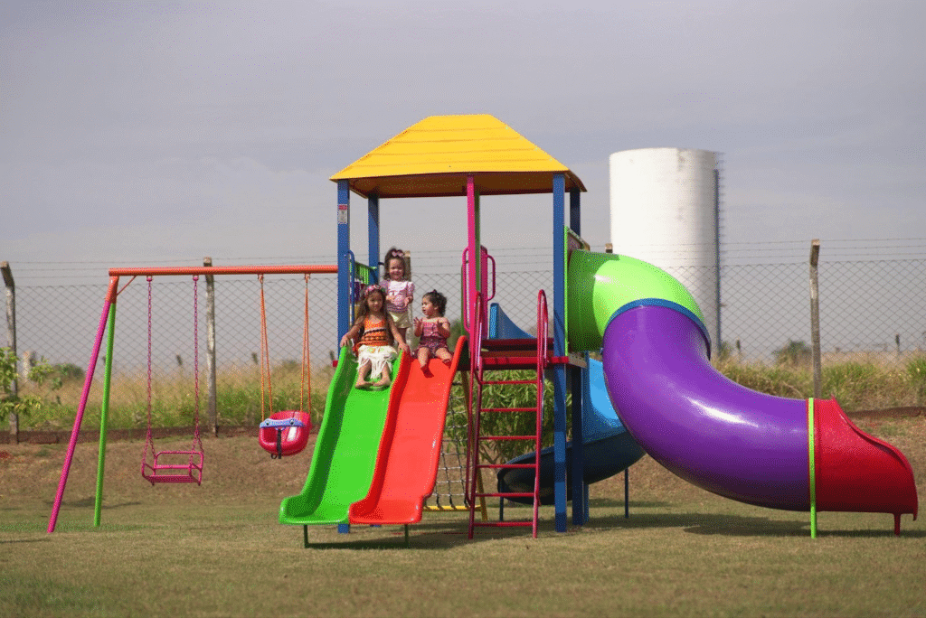 Crianças brincando em escorregador colorido em parque ao ar livre com balanços e estrutura de playground, ambiente ensolarado.