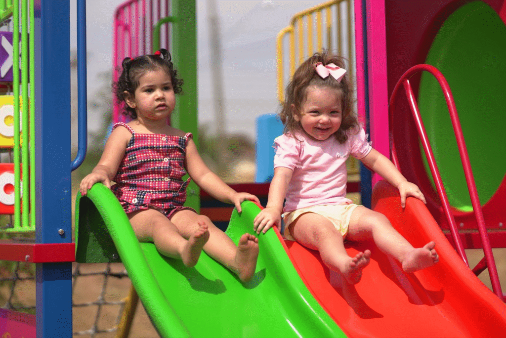 Duas meninas brincando em escorregadores coloridos em playground ao ar livre, uma com cabelo castanho escuro e a outra com cabelo castanho claro, sorrindo e se divertindo.