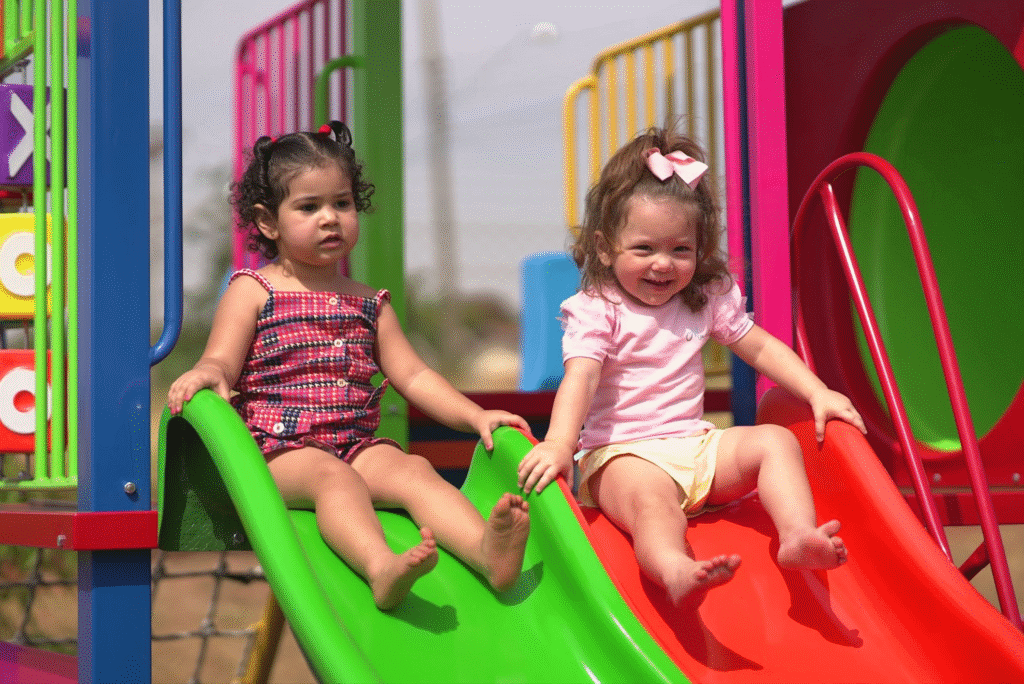 Duas crianças pequenas brincando juntas em um parque infantil com escorregadores verdes e vermelhos, sorrindo e se divertindo ao lado de cores vibrantes.