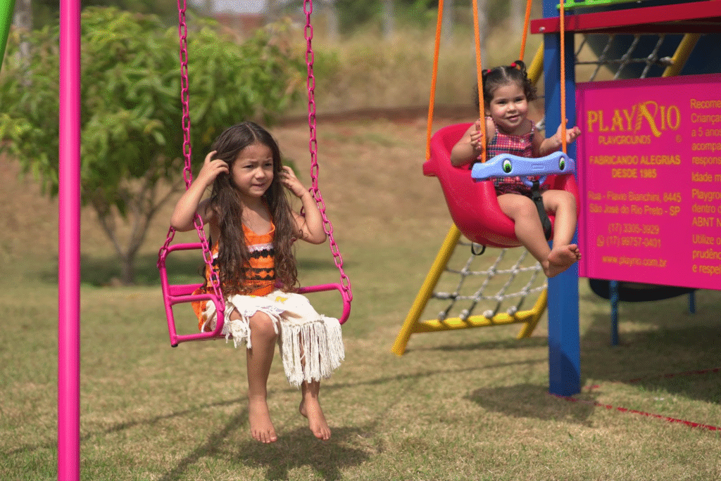 Duas meninas brincando no parquinho ao ar livre, uma na sanfona e outra na cadeira de balanço, sorrindo e se divertindo em um dia ensolarado.