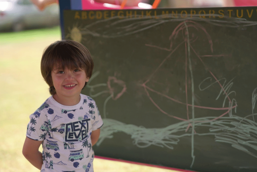 Criança sorrindo ao lado de um quadro-negro com desenhos coloridos de um barco à vela, durante aula ao ar livre na escola.