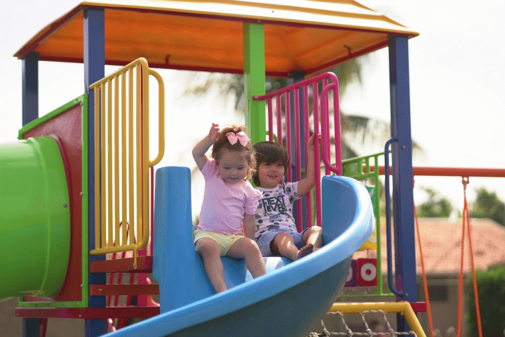 Duas crianças brincando em escorregador colorido na área de lazer ao ar livre, com fundo de árvores e céu nublado.