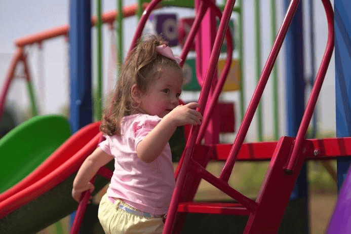 Menina pequena brincando em um parque de diversões com escadas e escorregador coloridos ao ar livre.