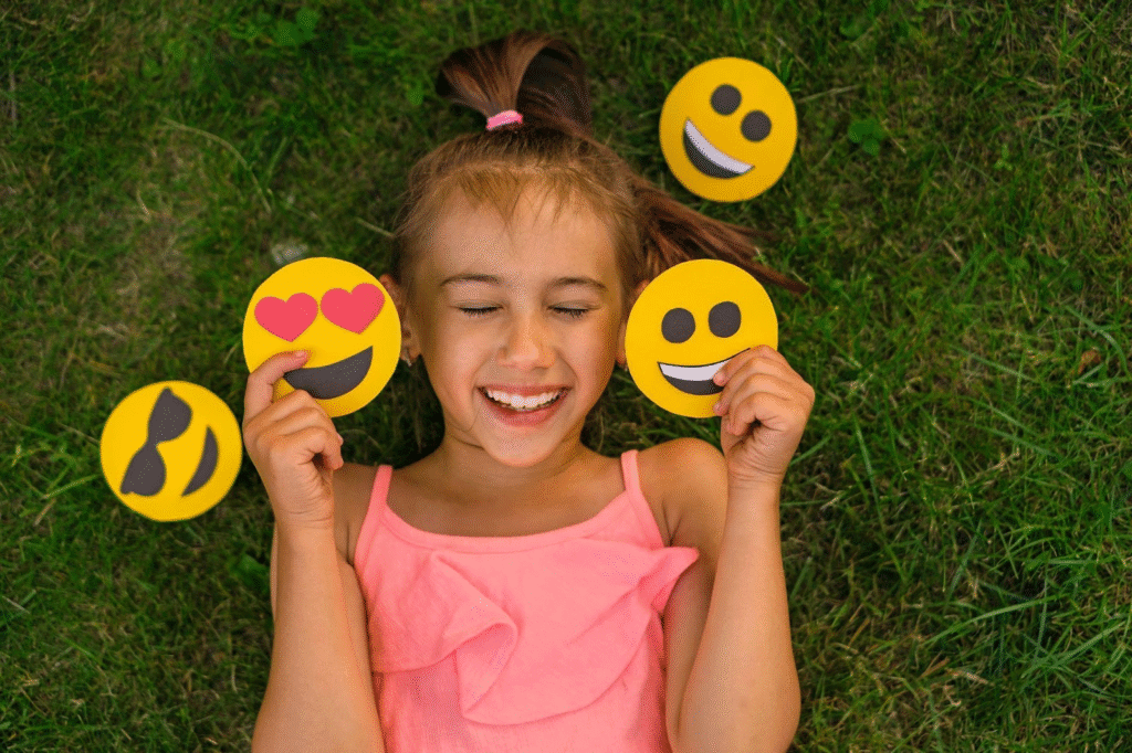 Menina sorrindo feliz enquanto deita na grama, rodeada por emojis de rostos sorridentes, apaixonados, de óculos escuros e com língua de fora, tema de emoções e expressões faciais, fundo natural verde.