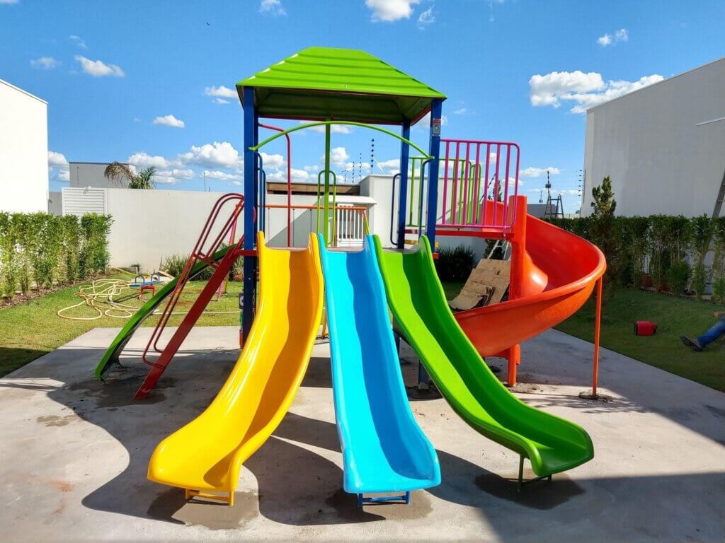 Parque infantil com escorregadores coloridos amarelo, azul, verde e um tobogã de tubo laranja, com céu azul e nuvens, ideal para diversão de crianças em área externa.